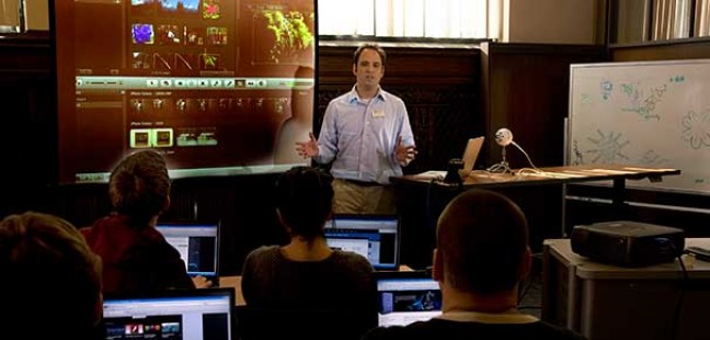 main lecturing a class with a computer screen projected behind him